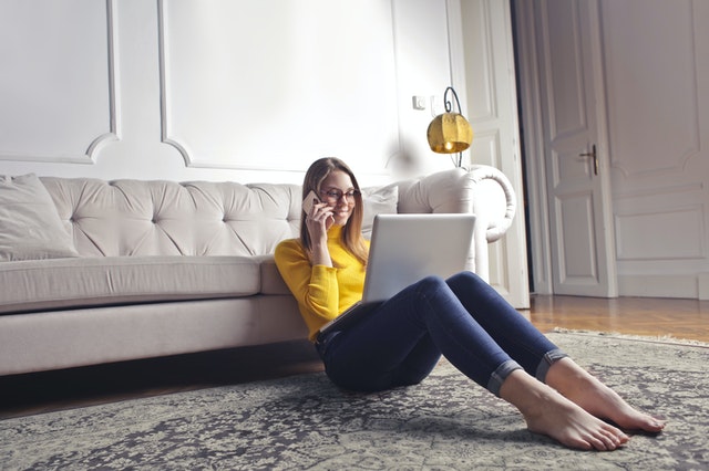 Woman in yellow shirt and jeans sitting on a rug with her back against a couch. She has a laptop on her lap and she is making a phone call to book an end of lease carpet cleaning services