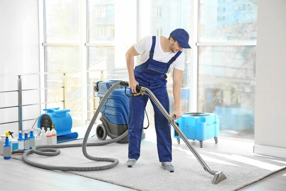 Man steam cleaning a rug with a machine. Cleaning product at the back an a fan to circulate air.